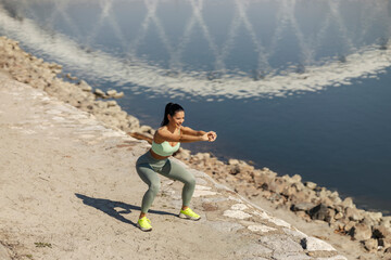 Full length of sportswoman in shape doing squatting endurance near river on riverbank.