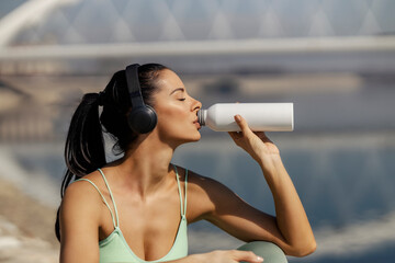 Side view of thirsty sportswoman sitting on riverbank, drinking fresh water from a bottle and...