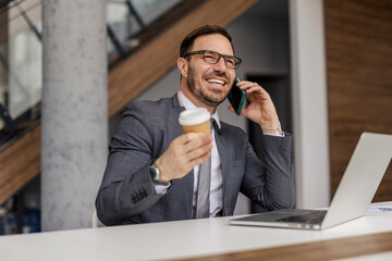 Friendly professional manager sitting at office with takeaway coffee in hands and having phone...