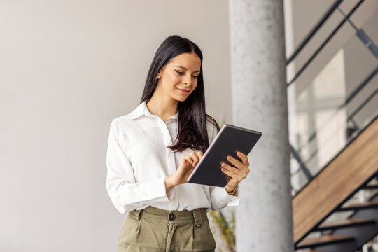 Portrait of busy female manager standing at corporate firm and scrolling on a tablet.