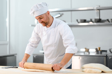 While working in bakery using rolling pin, male cook makes thin flat cakes from dough with rolling...