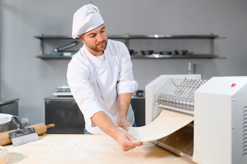 Traditional italian food. Side view of handsome chef rolling a dough through pasta machine in the kitchen