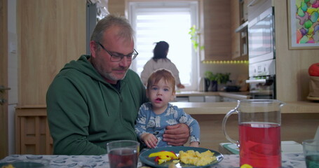 Father holding baby during family meal, baby gazing upwards with curiosity while sitting at dining...