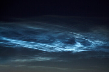 Rare noctilucent clouds glow with iridescent blue and white streaks across the darkening sky,...