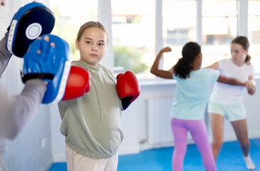 Concentrated sporty tween girl in boxing gloves practicing self-defense techniques in gym, throwing...