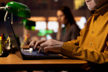 Aged man in a public library preparing thesis paperwork under a green lamp, surrounded by textbooks and study materials. Old pupil reflecting ambition for learning and achievements.