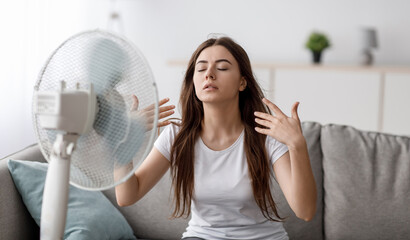 Sad young european woman suffering from heat sits on sofa, catches cold air from fan and waves...