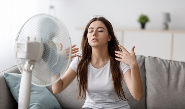 Sad young european woman suffering from heat sits on sofa, catches cold air from fan and waves hands to herself in living room interior. Summer weather and overheating without air conditioning at home