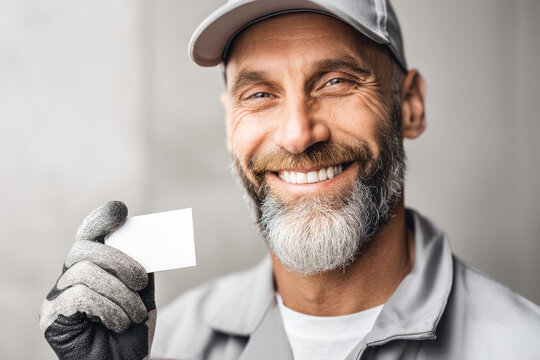An experienced smiling worker holding a blank business card, wearing work clothes and gloves, ready to offer professional services with confidence and approachability. - Powered by Adobe