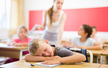 Portrait of tired bored small school boy lying and sleeping at desk in classroom during lesson
