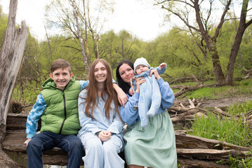 Family photo. Mother with three children pose for a photo, sitting on a log near a river in a park in spring. Teenagers sister and brother with a baby brother and mother.