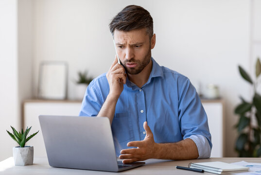 Worried Male Entrepreneur Having Problems With Laptop Computer At Work, Talking With Customer Support On Cellphone, Concerned Man Suffering Bad Internet Connection At Workplace In Office, Free Space