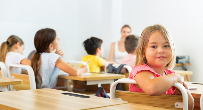 Portrait of cheerful cute blonde tween schoolgirl sitting at school desk at lesson in class, smiling looking at camera..