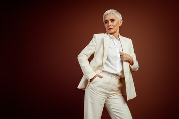 Elegant mature businesswoman in elegant white suit posing stylishly against a brown studio...