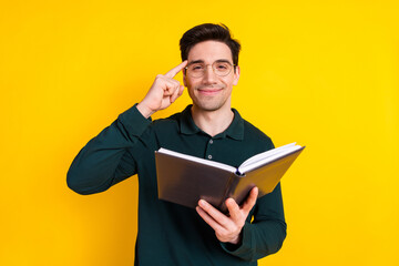 Young man in green shirt with glasses holding book and making thinking gesture against vibrant yellow background