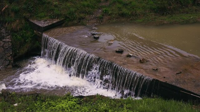 Water cascading over a small, concrete dam, creating a mini-waterfall effect. A waterfall, a stream of a cold spring water, showcasing natural beauty.