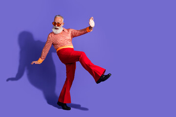 Obraz premium Elderly stylish man dancing with charisma in retro fashion, red flared pants, and bold vintage shirt on vibrant purple backdrop