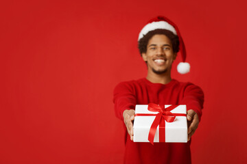 New Year presents and winter holidays. Focus on box with gift with ribbon in hands of african american man in sweater and santa claus hat, isolated on red background, copy space, studio shot