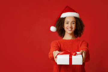 Merry Christmas and happy New Year. Cheerful young african american female in Santa Claus hat and sweater gives gift and looks at camera, isolated on red background, studio shot, empty space