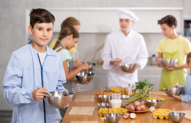 Teen boy participant in culinary master class holds dishes with ready-made successful pancake dough in hands. In background, blurry children stand near kitchen table and listen to chefs explanations