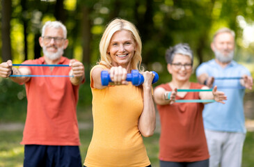 Active Lifestyle. Group of healthy senior people in sportswear training outdoors together, smiling elderly friends exercising with dumbbells and resistance band outside, doing fitness workout in park