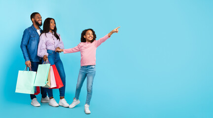 Family Shopping Concept. Full length body size portrait of African American cheerful people holding shopping bags with new clothes, excited little girl pointing aside at copy space, blue studio wall © Prostock-studio