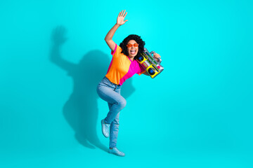 Cheerful young woman in colorful outfit with boom box, dancing on vibrant turquoise background,...