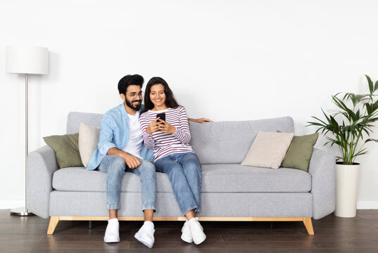 Relaxed cheerful beautiful young multicultural couple middle eastern guy and indian woman sitting on sofa at home, using smartphone and smiling, shopping online, surfing on Internet, copy space