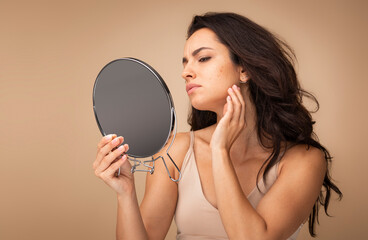 Stressed sad brunette young woman wearing beige top looking at mirror and touching her face over beige studio background, having skin problems. Acne, pimples, wrinkles, dull skin