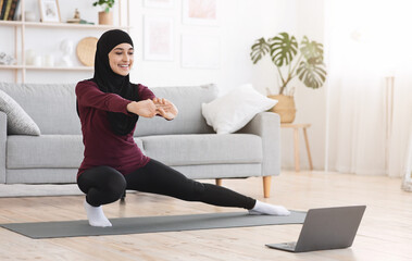 Happy arabic woman in hijab doing lunges in front of laptop at home, making fitness training via...