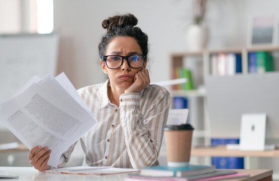 Tired female office worker looking at camera and holding documents, sitting at desk in office. Unhappy woman feeling stressed because of lot of paperwork