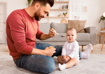 Cute Young Dad Spoon Feeding Little Baby Toddler Caring For Daughter During Paternity Leave Sitting On Floor At Home. Healthy Nutrition For Newborn, Fatherhood And Child Care Concept