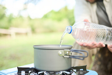 Unrecognizable person pouring drinking water into a pot as part of the process of cooking outdoors, in a portable gas stove, while camping. Selective focus on water. Close up view, with copy space.