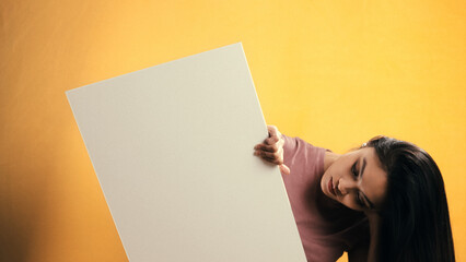 Studio shot of young Asian woman holding a white blank board with copy space on a yellow background