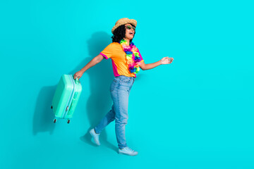 Joyful young woman in casual summer attire walking with a suitcase against a vibrant turquoise...