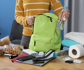 Survival kit. Woman packing different emergency supplies into backpack at wooden table indoors,...