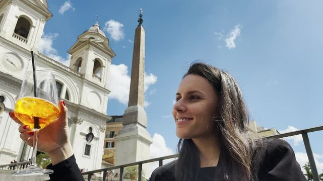 Businesswoman toasting with refreshing orange cocktail cheering against amazing Spanish staps, obelisk and church of Trinita dei Monti at background, relaxing and enjoying the view of ancient Rome