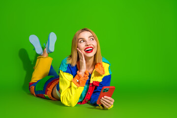 Young cheerful woman in vibrant colorful outfit holding smartphone while lying on green background, embodying fun and expressive style.