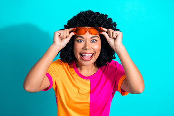 Joyful woman in colorful outfit holding sunglass with a big smile against a bright turquoise background