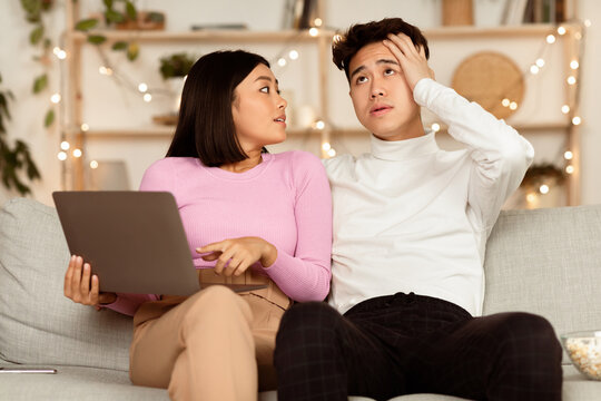 Asian Shopaholic Wife Shopping Online With Shocked Husband, Asking Him Buy Expensive Clothes And Spend Too Much Money Sitting On Sofa At Home. Prodigality, Shopaholism And Bad Spending Habits Concept