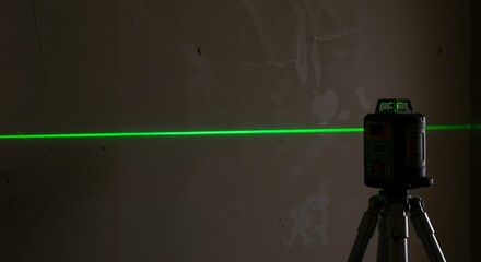 A green laser level mounted on a tripod projects a bright, precise horizontal line onto an unfinished wall in a dimly lit room for construction or renovation purposes.