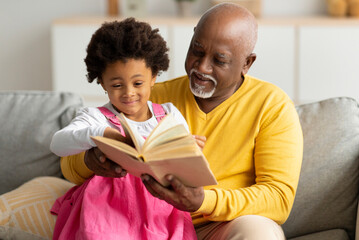 Cheerful cute african american little granddaughter and senior grandpa reading book with fairy...