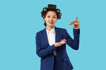 Young Asian stewardess with hair curlers and wooden airplane on blue background
