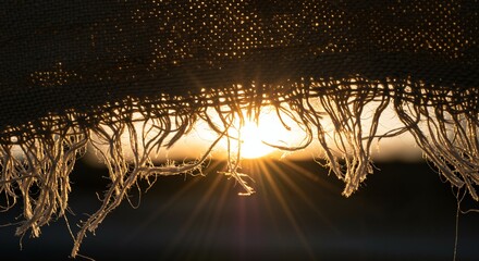 Golden hour sunlight streams through the tattered, frayed edge of a dark, textured fabric, creating a dramatic silhouette and a starburst effect as the sun peeks through the loose threads.