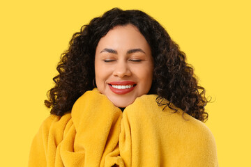 Beautiful young African-American woman with cozy blanket on yellow background