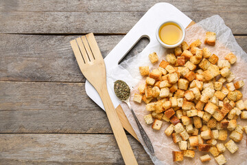 Cutting board with crunchy croutons and bowl of oil on wooden background