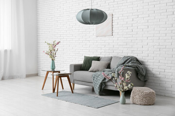 Sofa with coffee tables and vases of blooming branches near white brick wall in living room