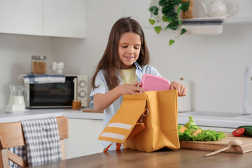 Cute girl packing school lunch box into bag in kitchen