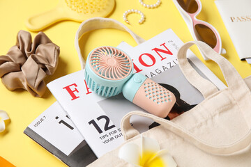 Bag with female summer accessories, fashion magazines and mini fan on yellow background