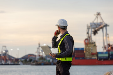 A male port engineer wearing a safety vest and helmet holds a laptop and talks on a radio at a busy shipping dock, overseeing cargo operations with cranes and containers in the background.